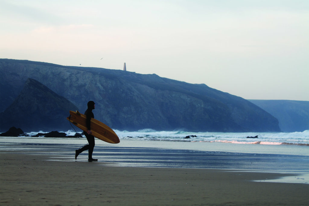 James Otter walking out to the surf under the cliffs at Porthtowan with his fetch wooden surfboard.
