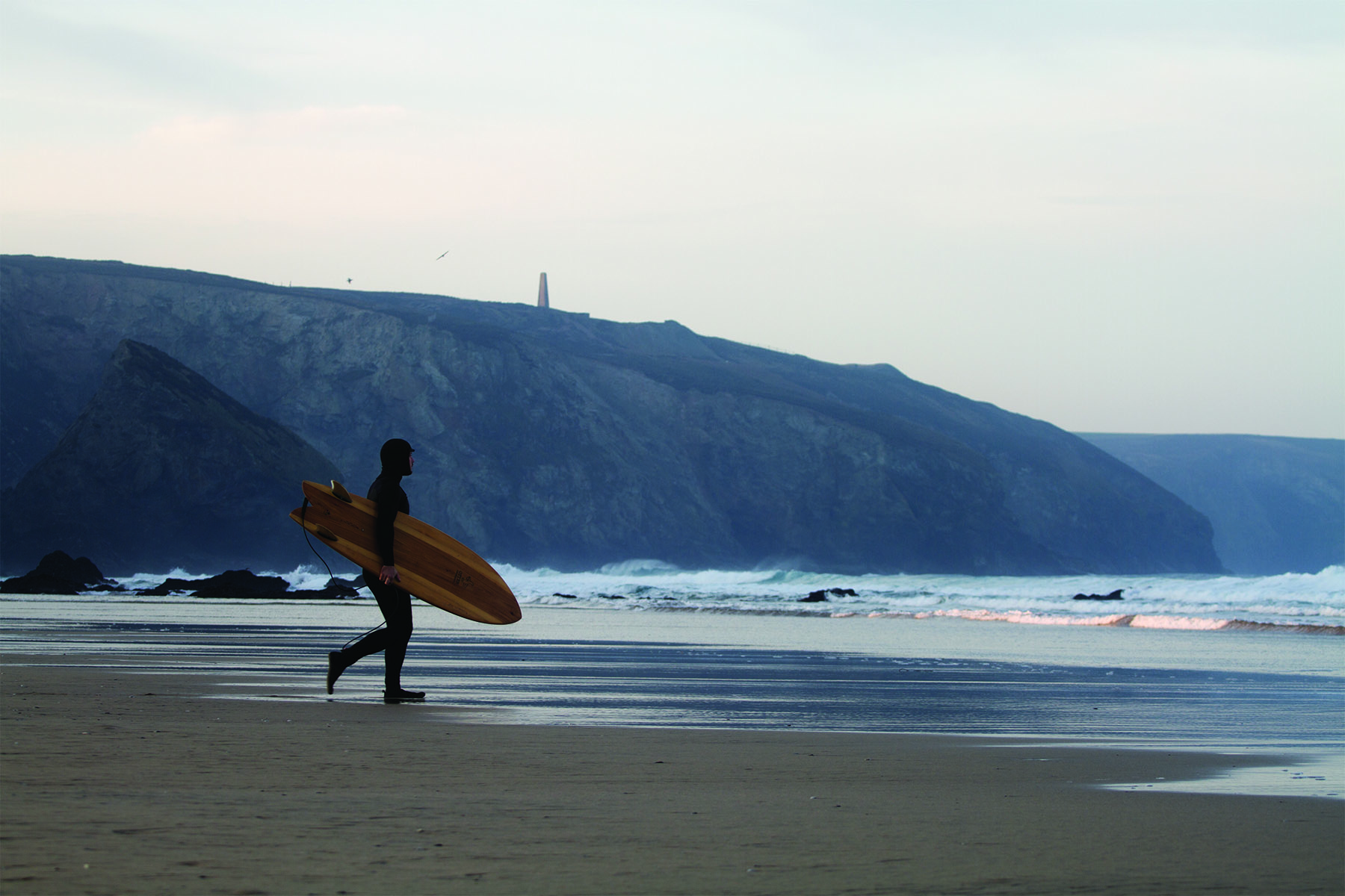 James Otter walking out to the surf under the cliffs at Porthtowan with his fetch wooden surfboard.