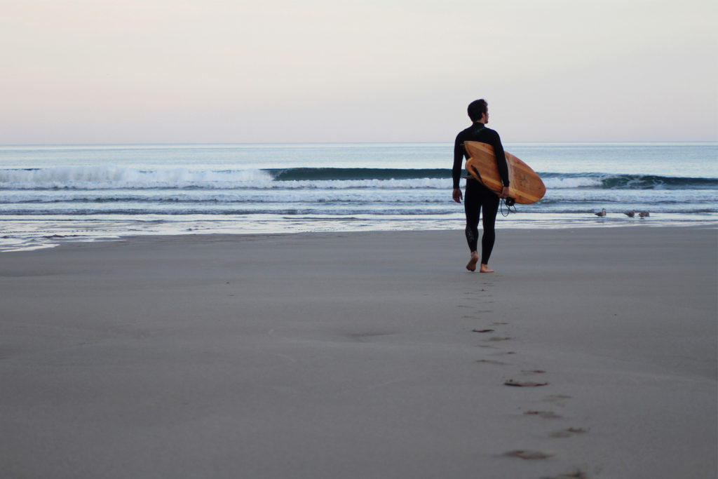 James Otter walking out to the surf at Chapel Porth, Cornwall with his fetch wooden surfboard.