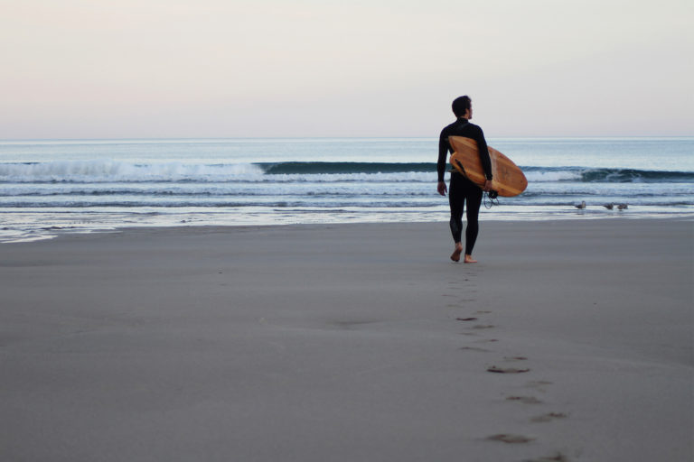 James Otter walking out to the surf at Chapel Porth, Cornwall with his fetch wooden surfboard.