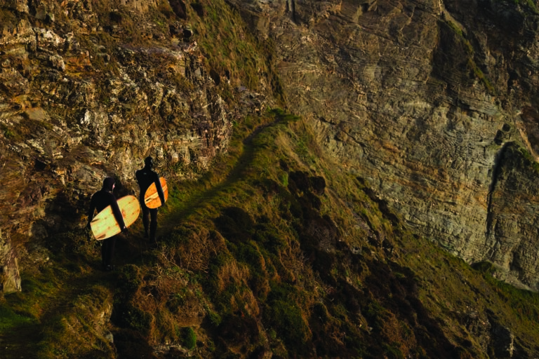 Tony and James Otter walk down the cliff path with their wooden surfboards. Jetty and Seadar