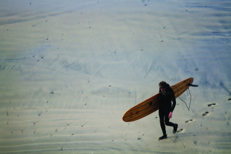 Mat Arney walks back across the sand at Gwithian with his wooden Otter Surfboards Pier.