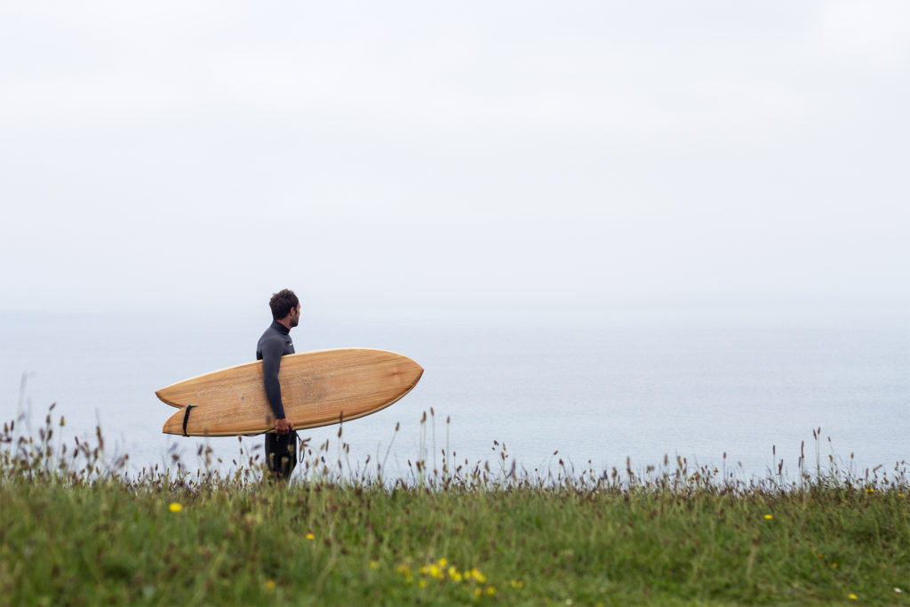 Ally Stands on the cliffs in the grassy meadow at Porthtowan with his Otter Wooden surfboard Woodburner.