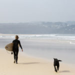 Sophie Hellyer walks along a sandy beach in the wind at st Ives bay with buddy the dog and her otter surfboards wooden surfboard Coaster