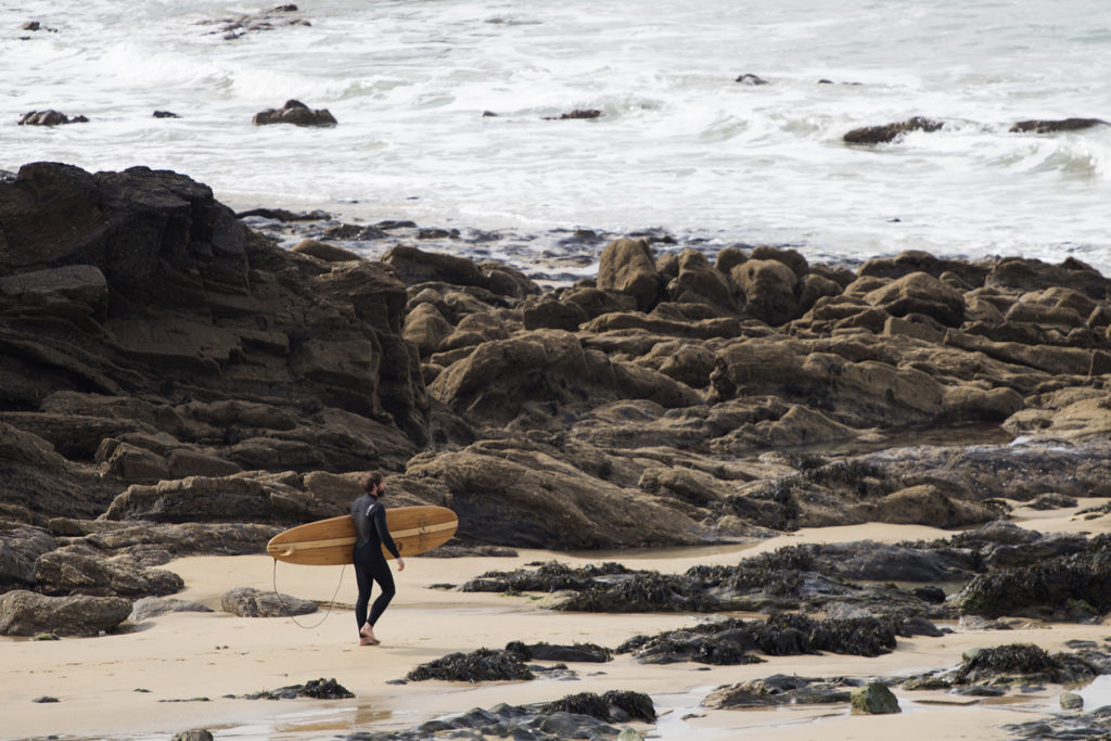 Island Hopper Tony walks into the surf at Church Cove with his wooden surfboard otter